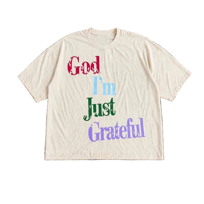 A cream-colored short-sleeve t-shirt with the phrase 'God I'm Just Grateful' printed in a multi-colored retro font, laid flat on a dark wood plank background.